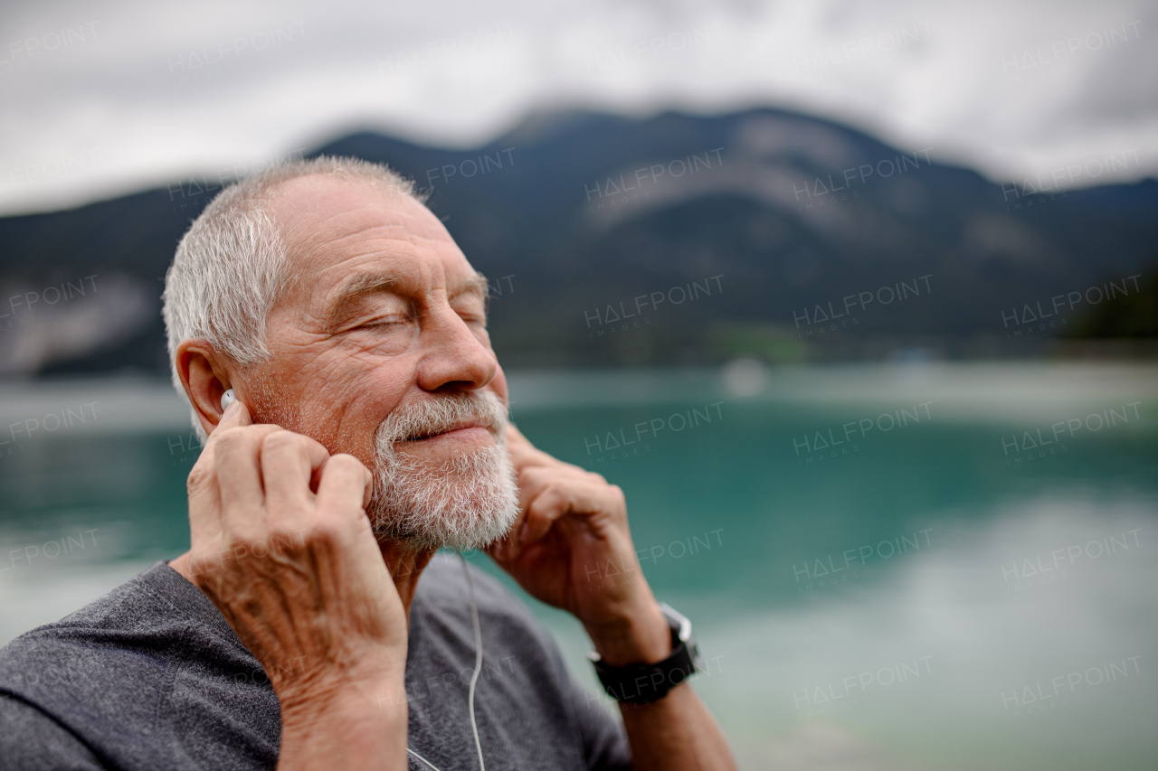 Senior listening music while running by lake in nature. Elderly man exercising to stay healthy, vital, enjoying physical activity, relaxation outdoors.