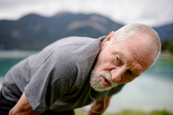 Senior breathing after run by lake in nature. Elderly man exercising to stay healthy, vital, enjoying physical activity, relaxation outdoors.