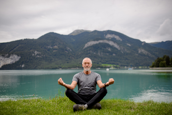 Senior man doing outdoor yoga, meditating by the lake in mountains. Elderly sportsman spending active vacation in nature. Pensioner stretching after outdoor workout