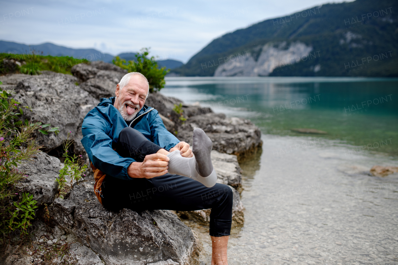 Active elderly man putting on sock after barefoot walk in cold water in mountain lake. Senior tourists enjoying nature during early spring day.