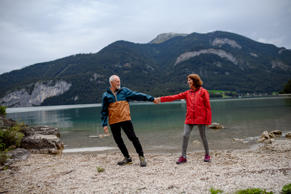 Active elderly couple hiking together in spring mountains, walking on pebble beach of mountain lake. Senior tourists enjoying peaceful nature.