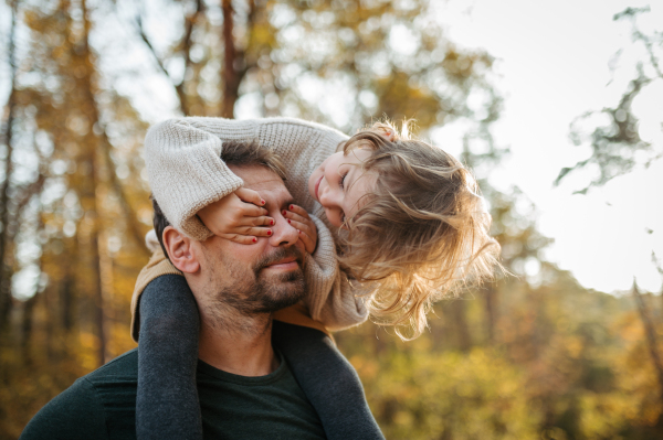 Father walking with daughter on his shoulders across autumn forest. Dad and girl having fun outdoors