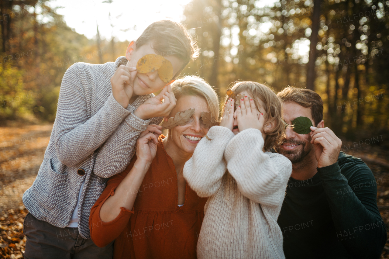 Portrait of family sitting in the middle autumn forest covering eyes with colorful leaves. Nuclear family spending weekend outdoors