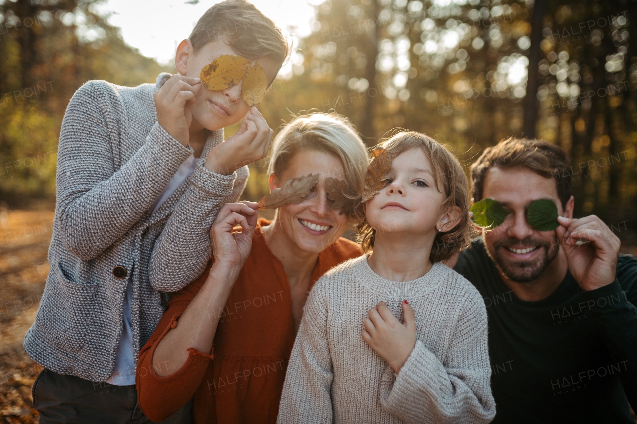 Portrait of family sitting in the middle autumn forest covering eyes with colorful leaves. Nuclear family spending weekend outdoors