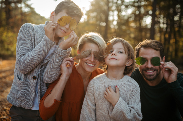 Portrait of family sitting in the middle autumn forest covering eyes with colorful leaves. Nuclear family spending weekend outdoors