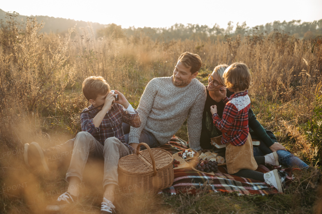 Portrait of family having picnic in autumn nature, eating sandwiches, fruits and cheese in the middle of meadow. Young boy taking pictures with vintage camera