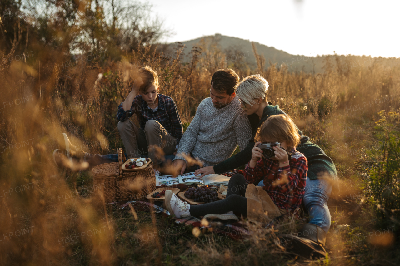 Portrait of family having picnic in autumn nature, eating homemade food in the middle of meadow. Young girl taking pictures with vintage camera