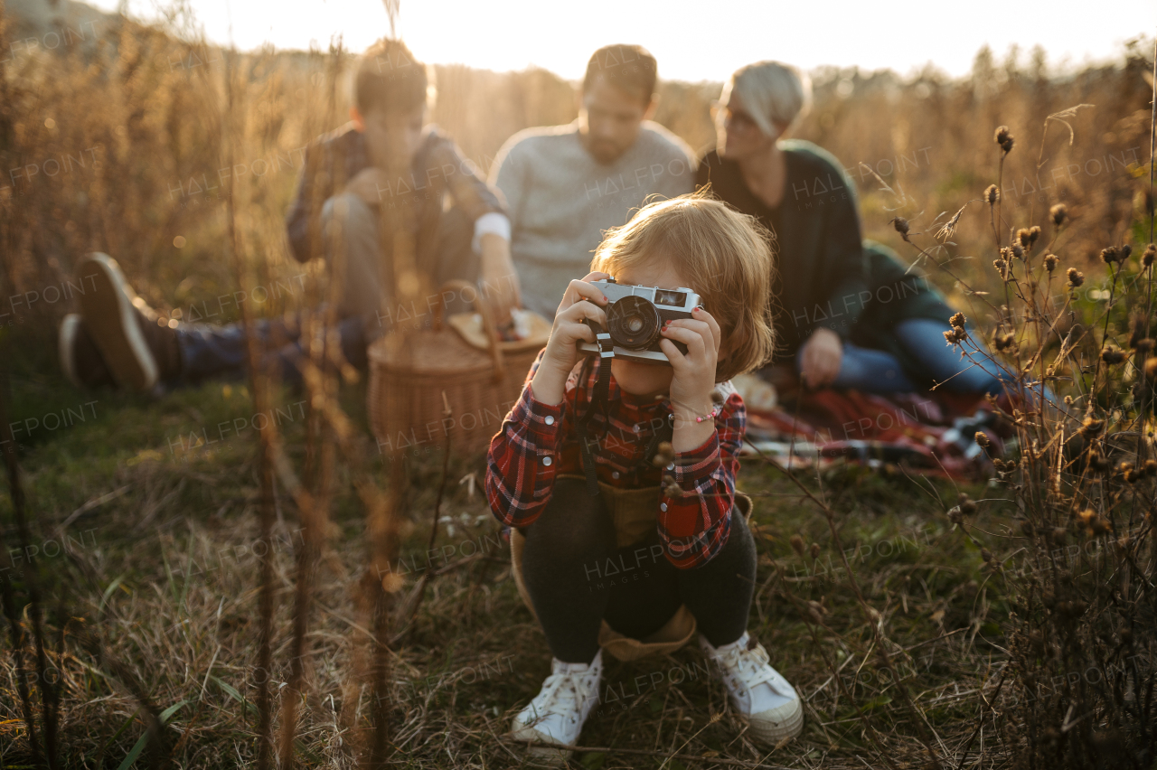 Portrait of family having picnic in autumn nature, eating homemade food in the middle of meadow. Young girl taking pictures with vintage camera