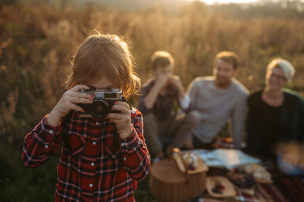 Portrait of family having picnic in autumn nature, eating fruits and cheese in the middle of meadow. Young girl taking pictures with vintage camera