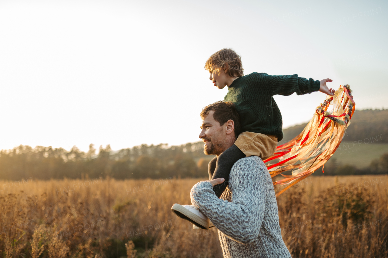 Father walking with daughter on his shoulders across autumn meadow. Girl holding wreath with ribbons