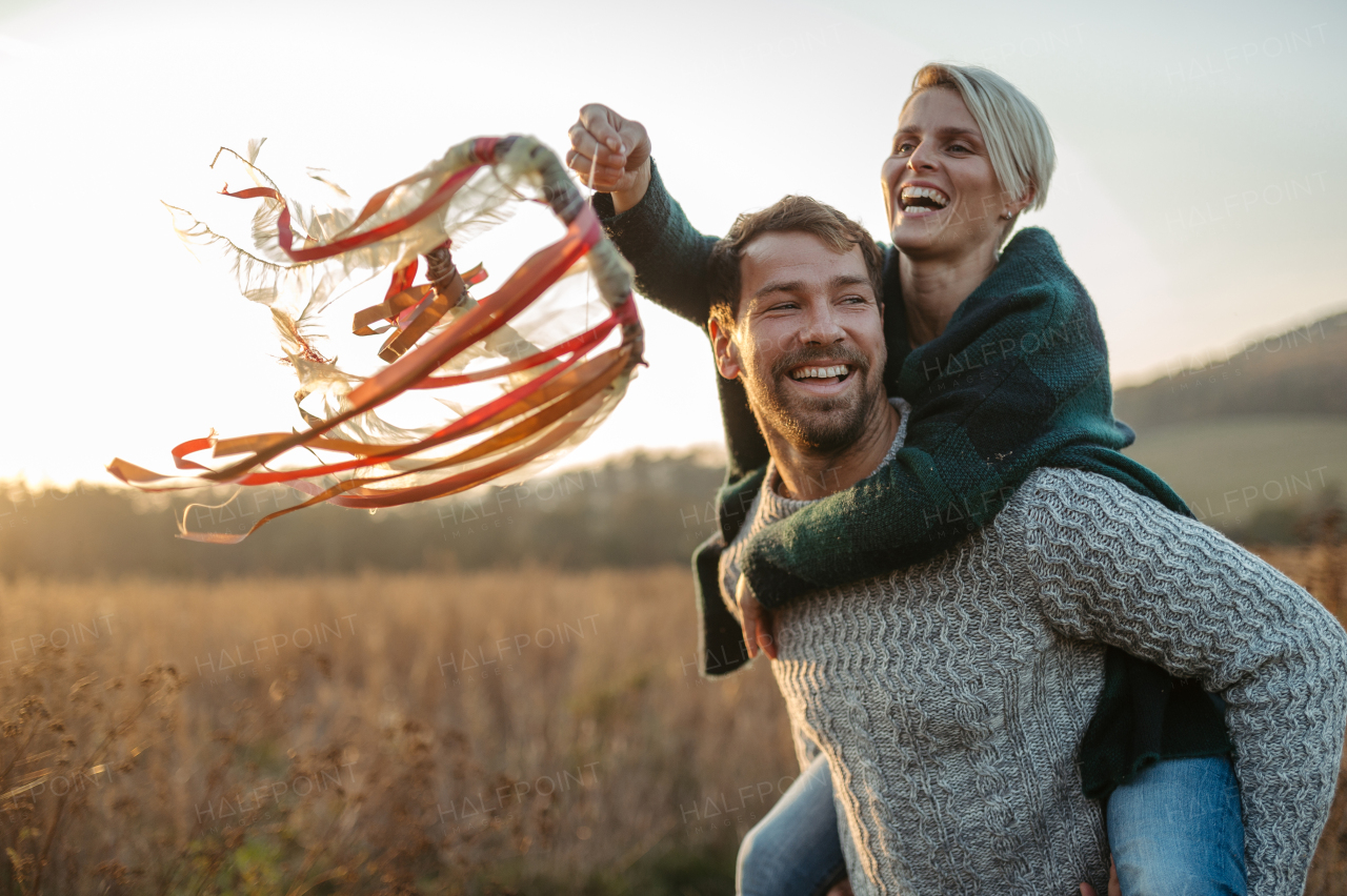 Couple in love on a walk in autumn forest. Husband piggycarrying wife on back, holding wreath with ribbons