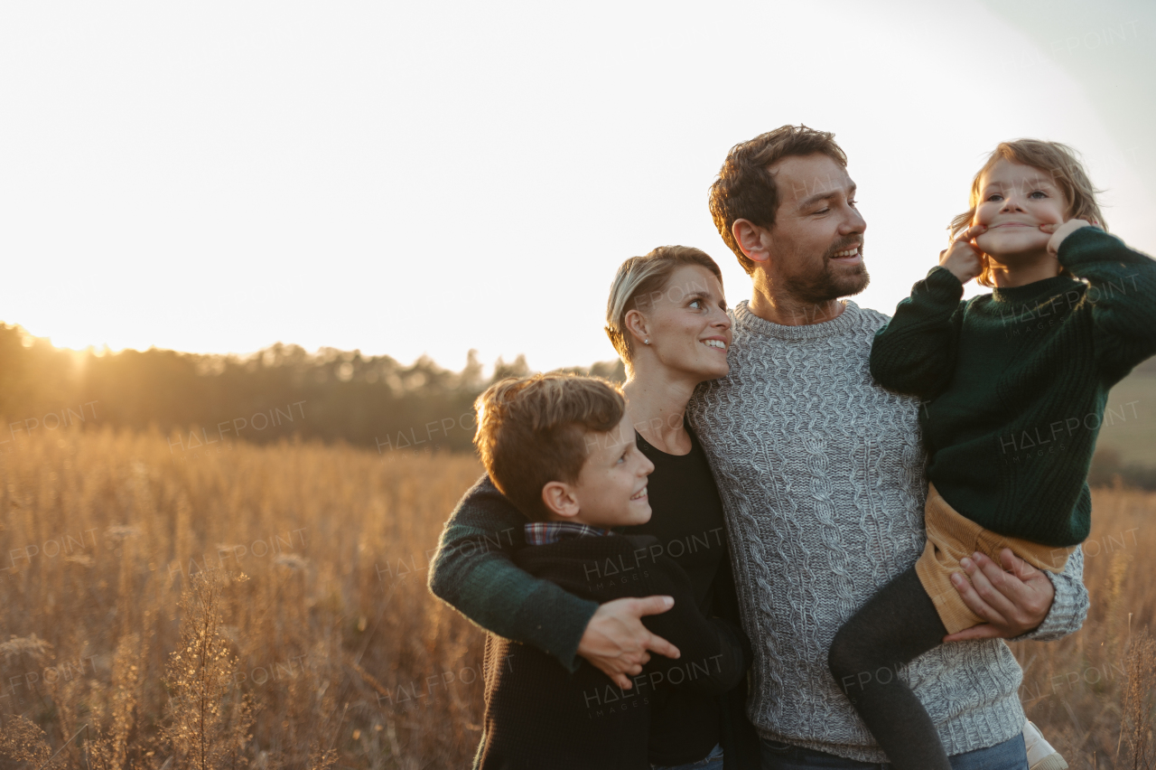 Portrait of young family with two kids on a walk in autumn nature, standing in the middle meadow during sunset.