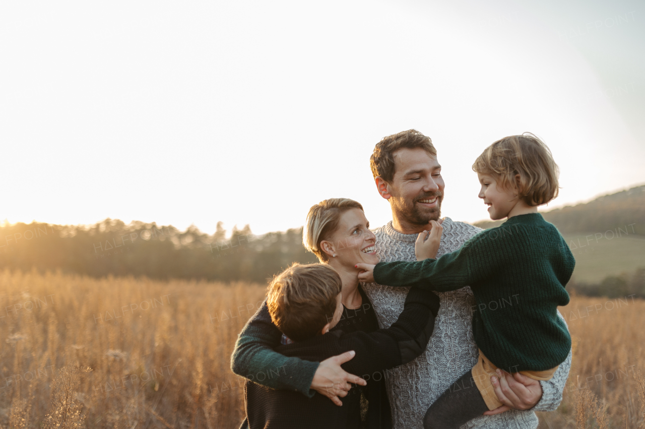 Portrait of young family with two kids on a walk in autumn nature, standing in the middle meadow during sunset.