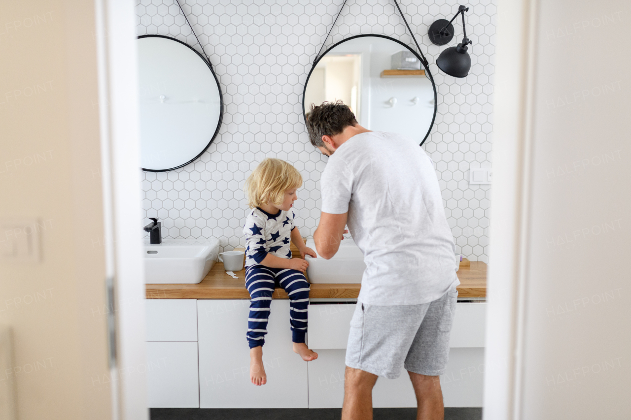 Father brushing son's teeth, boy sitting on bathroom countertop. Morning routine for young boy before going to preschool.