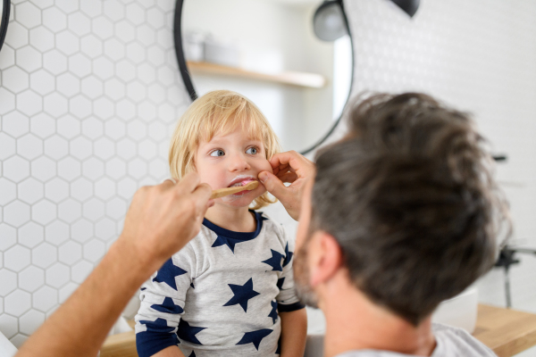 Father brushing son's teeth, boy sitting on bathroom countertop. Morning routine for young boy before going to preschool.