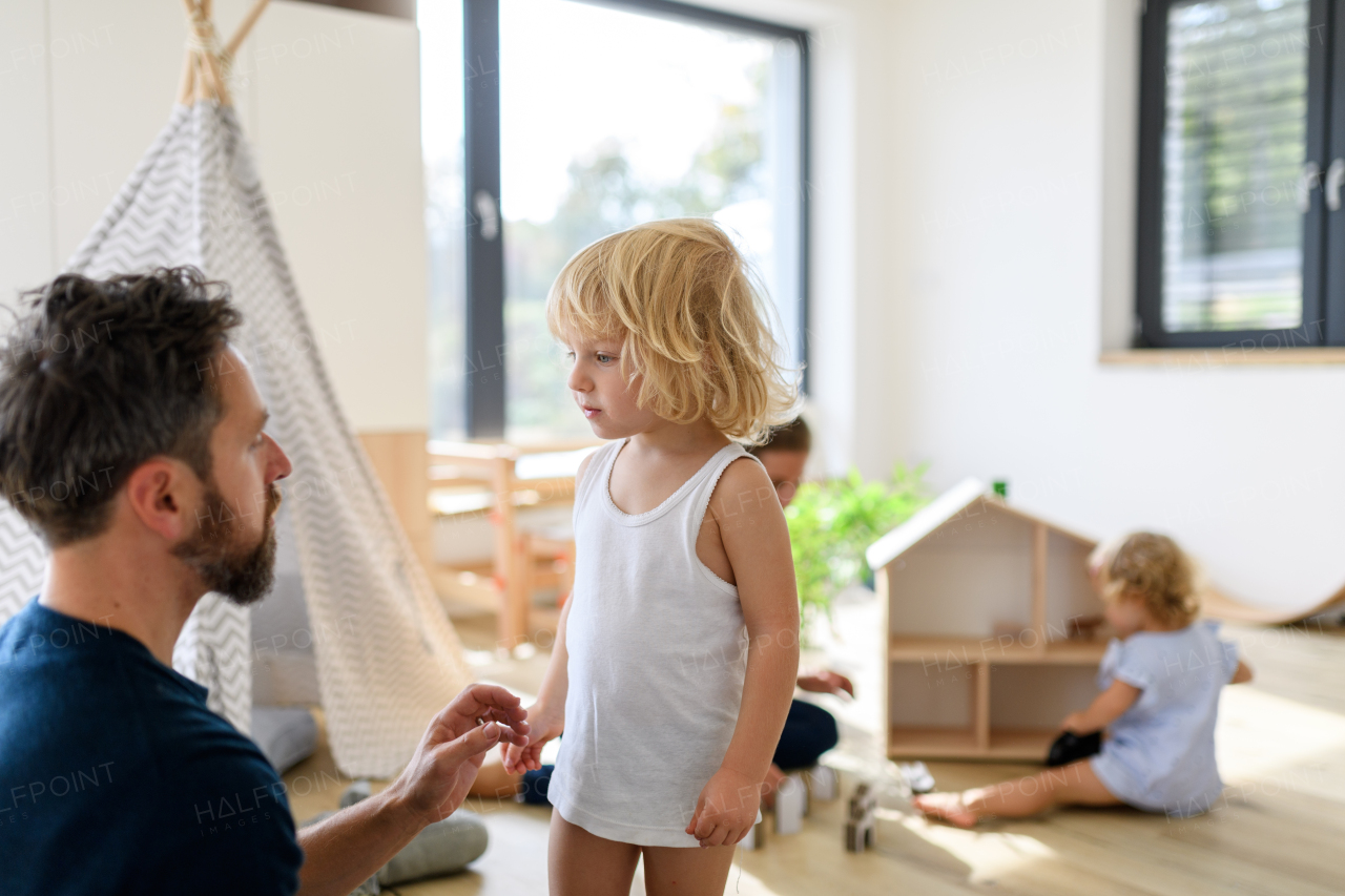 Father putting clothes on small boy, who trying to run off. Young nuclear family having fun at home, spending weekend day indoors.