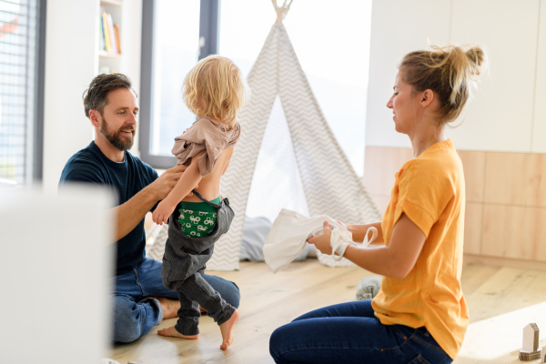 Parents putting clothes on small boy, who trying to run off. Young nuclear family having fun at home, spending weekend day indoors.