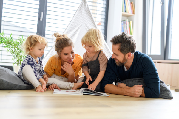 Young nuclear family playing with toys in a living room. Parents and children lying on floor, looking at children's story, picture book, spending weekend day indoors.