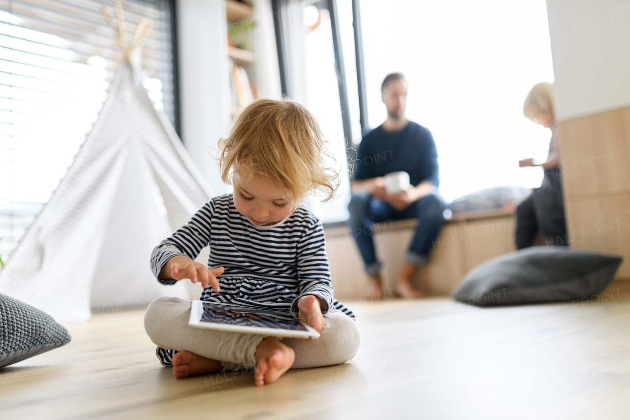 Cute little girl is sitting on the ground, scrolling on a tablet.