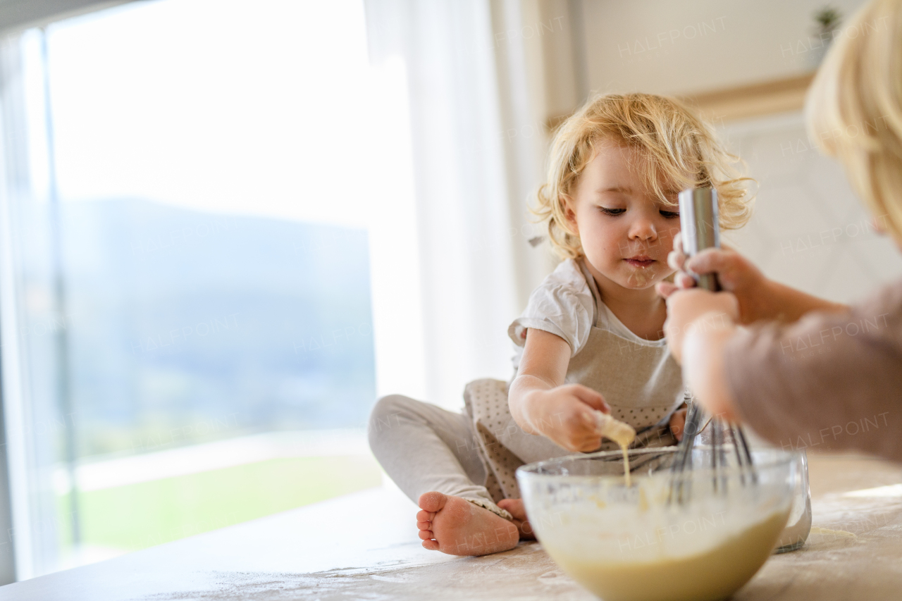 Little cute girl sitting on kitchen countertop, tasting pancake batter with finger. Nuclear family spending weekend day indoors, cooking together.