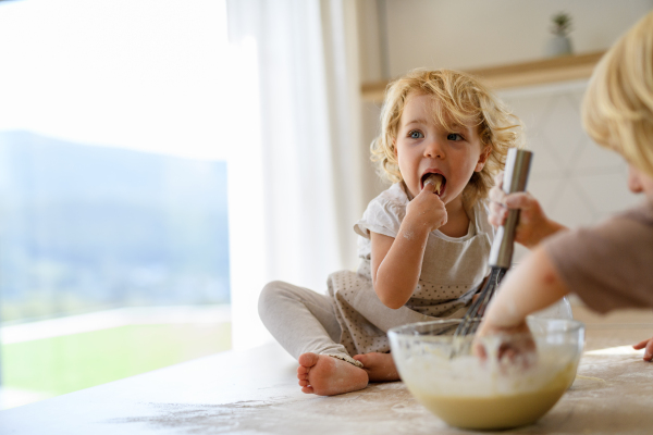 Little cute girl sitting on kitchen countertop, tasting pancake batter with finger. Nuclear family spending weekend day indoors, cooking together.