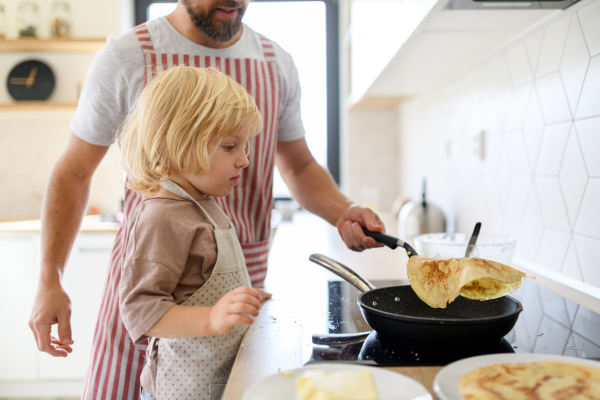 Young boy learning how to make pancakes, standing by stove. Parents and children in kitchen, preparing pancake batter, spending weekend day indoors