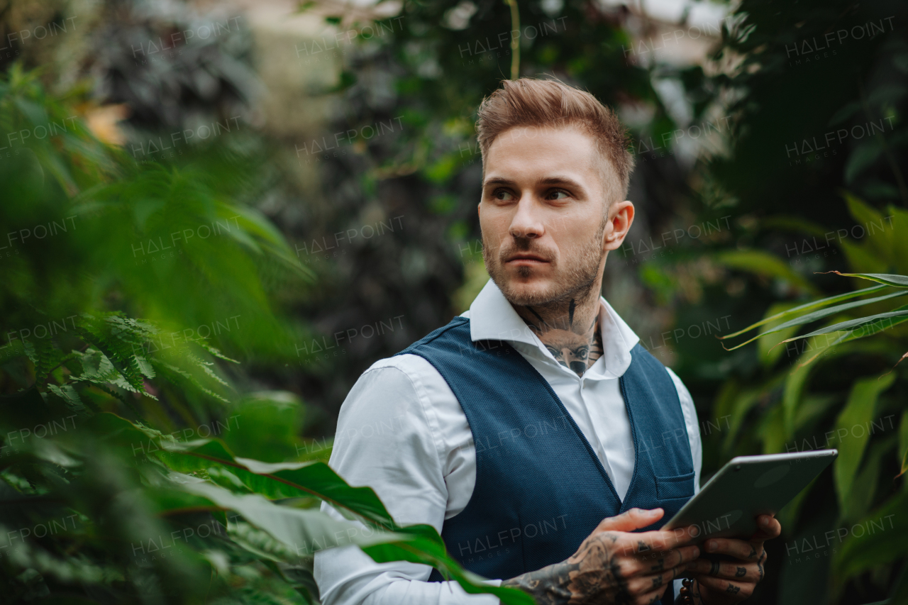 Businessman with tablet, standing in botanical garden, in the middle of lush green foliage. Concept of corporate social responsibility and ESG.