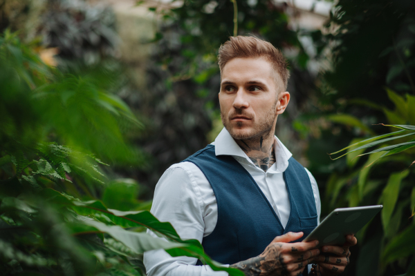 Businessman with tablet, standing in botanical garden, in the middle of lush green foliage. Concept of corporate social responsibility and ESG.