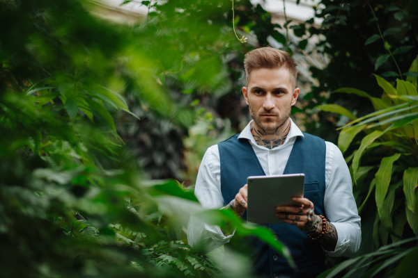 Businessman with tablet, standing in botanical garden, in the middle of lush green foliage, looking at camera. Concept of corporate social responsibility and ESG.