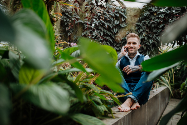 Businessman making phone call with smartphone, sitting in botanical garden, in the middle of lush green foliage.Concept of corporate social responsibility and ESG.