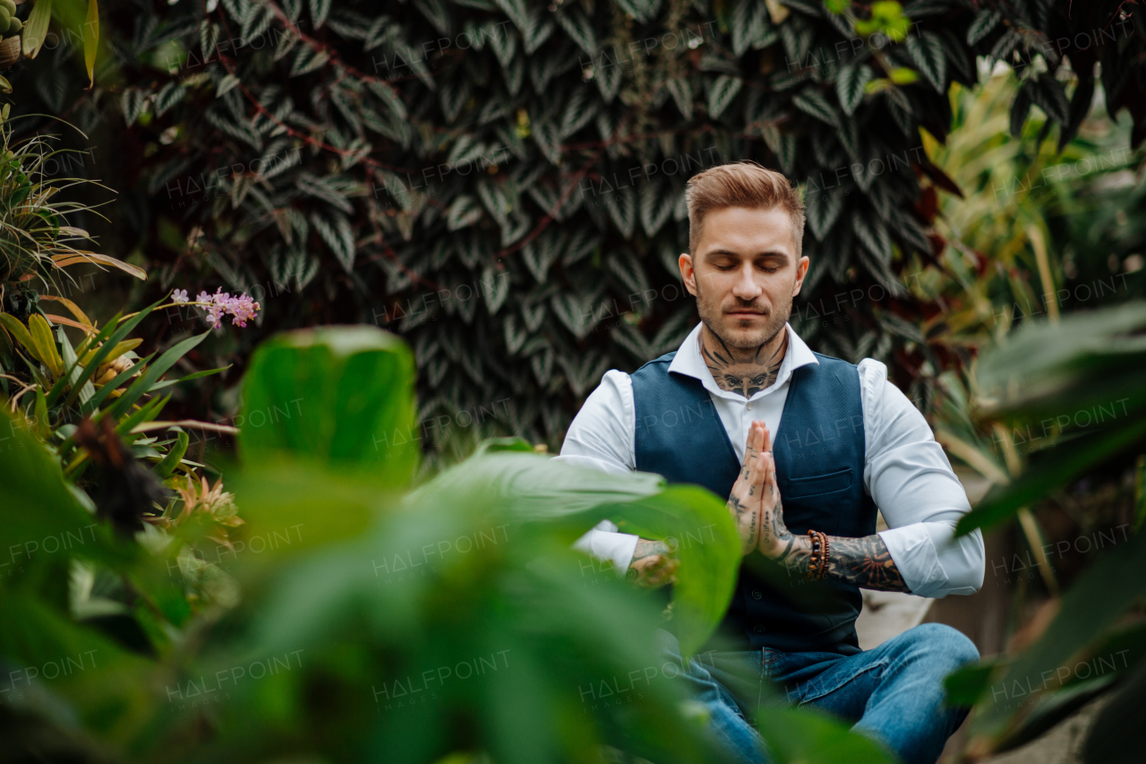 Man with tattoo meditating in botanical garden, in the middle of lush green foliage. Taking care of his mental health, relaxing.