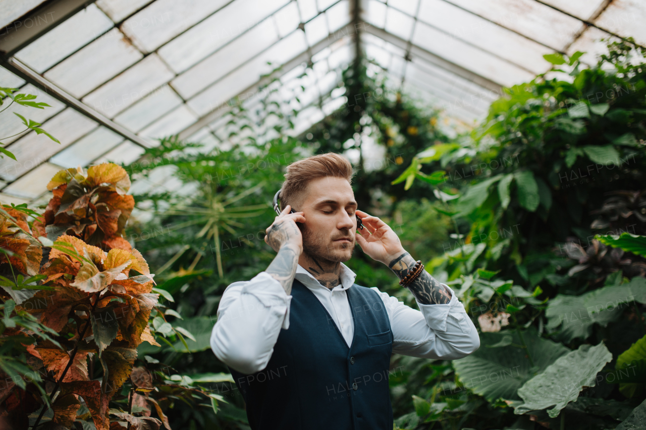 Man with tattoo listening meditation music in botanical garden, in the middle of lush green foliage. Taking care of his mental health, relaxing.