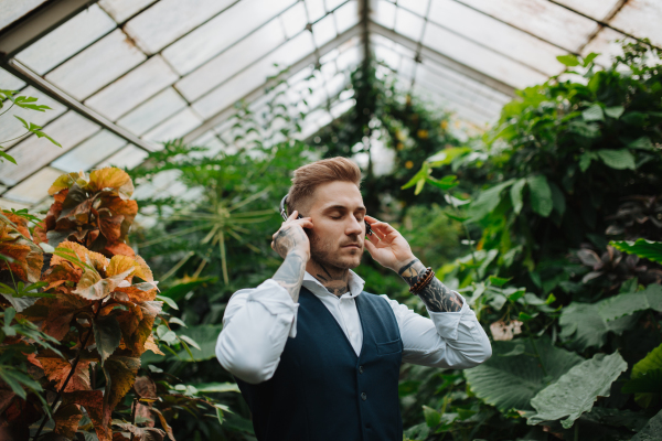 Man with tattoo listening meditation music in botanical garden, in the middle of lush green foliage. Taking care of his mental health, relaxing.