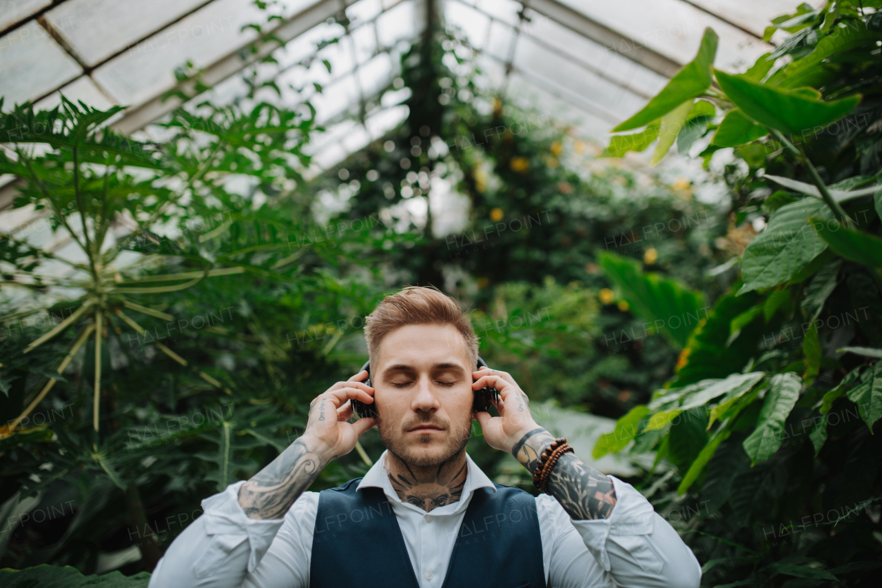 Man with tattoo listening meditation music in botanical garden, in the middle of lush green foliage. Taking care of his mental health, relaxing.