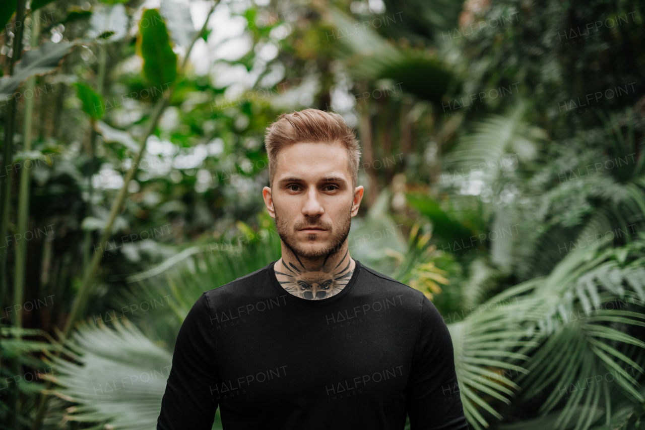 Handsome man with tattoo standing in botanical garden, in the middle of lush green foliage.