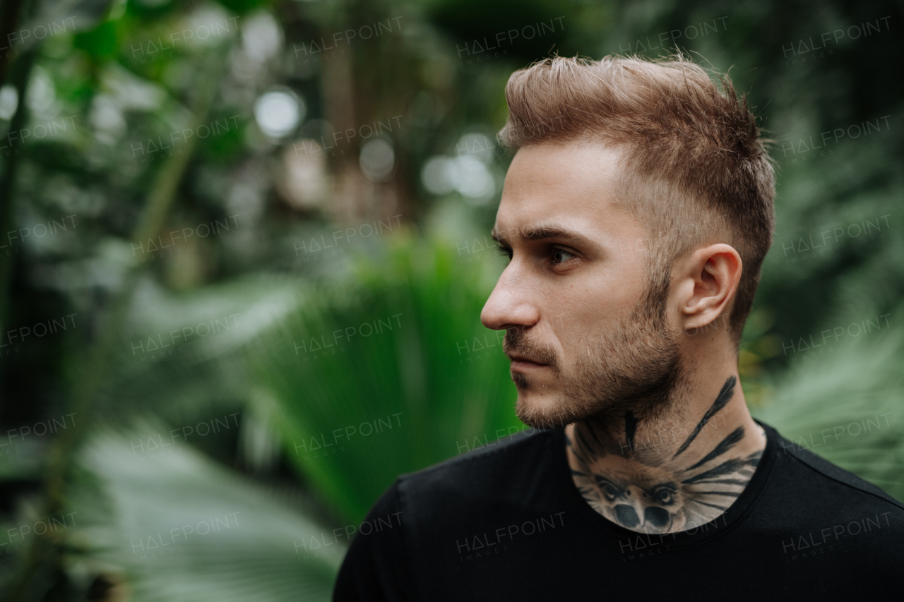 Handsome man with tattoo standing in a botanical garden, in the middle of lush green foliage.