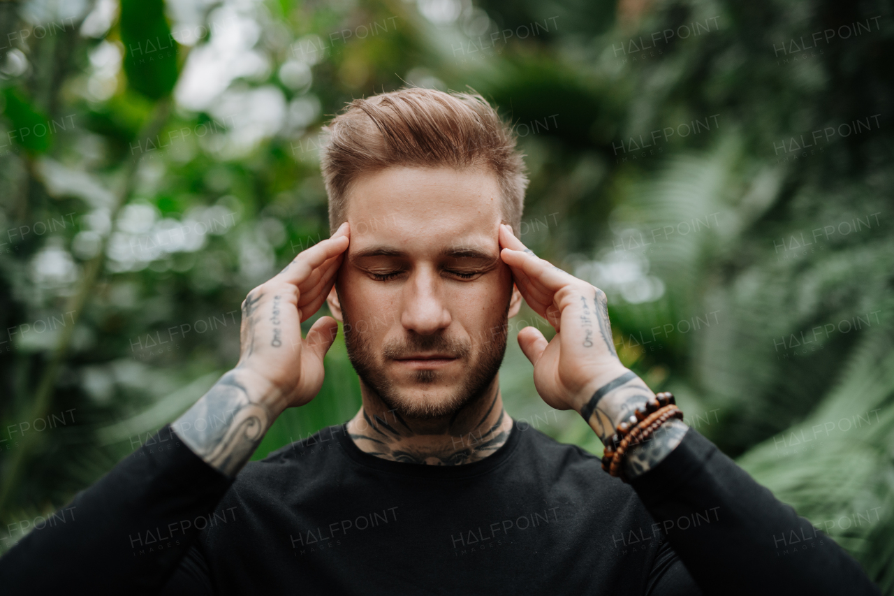 Man with tattoo standing in botanical garden, in the middle of lush green foliage, holding head in hands, feeling overwhlemed.