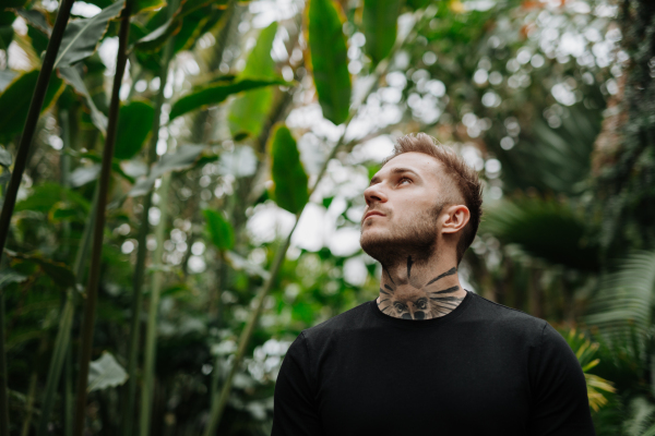 Handsome man with tattoo standing in botanical garden, in the middle of lush green foliage.