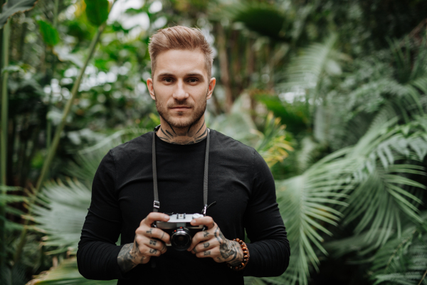 Man with tattoos standing in botanical garden, in the middle of lush green foliage, holding camera and taking photography.