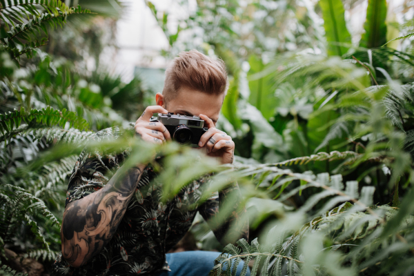 Man with tattoos standing in botanical garden, in the middle of lush green foliage, holding camera and taking photography.