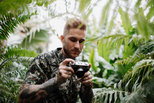 Man with tattoos standing in botanical garden, in the middle of lush green foliage, holding camera and taking photography.