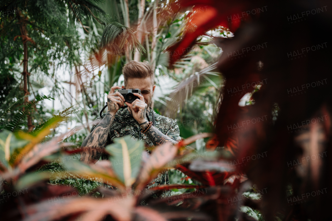 Man with tattoos standing in botanical garden, in the middle of lush green foliage, holding camera and taking photography.