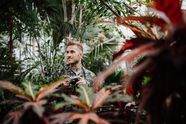 Man with tattoos standing in botanical garden, in the middle of lush green foliage, holding camera and taking photography.