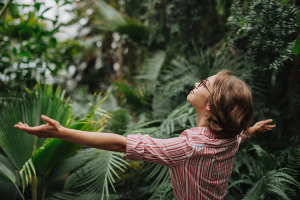 Woman standing in botanical garden, in the middle of lush green foliage, arms open. Rear view.