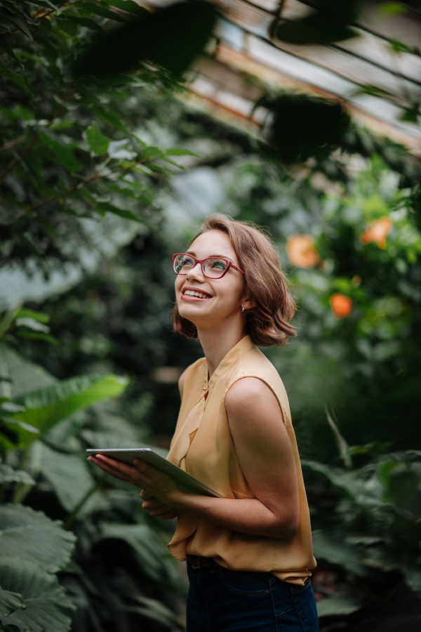 Businesswoman with tablet, standing in botanical garden, in the middle of lush green foliage, looking at camera. Concept of corporate social responsibility and ESG.