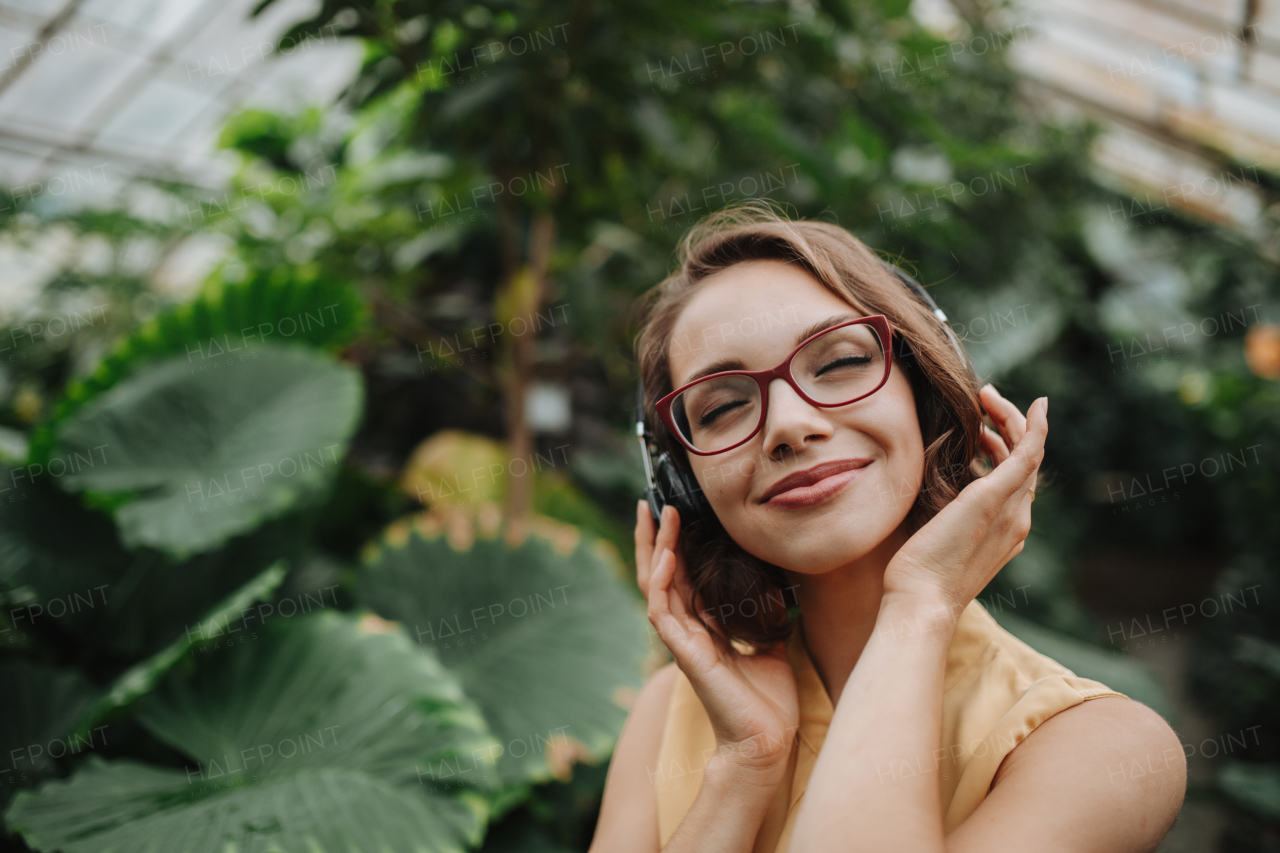 Beautiful woman standing in botanical garden, in the middle of lush green foliage with headphones on head, listening and enjoying music.