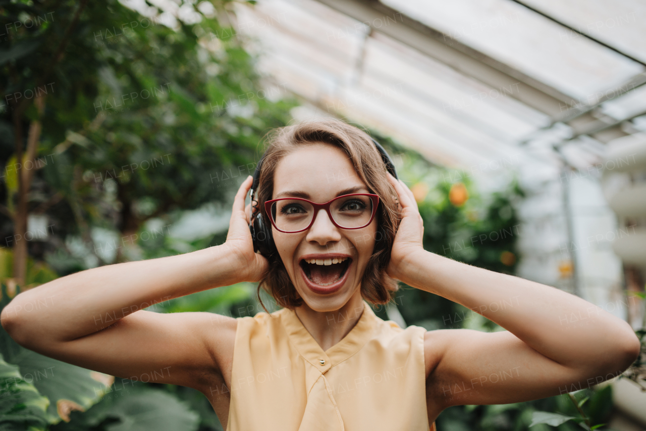 Beautiful woman standing in botanical garden, in the middle of lush green foliage with headphones on head, listening and enjoying music.