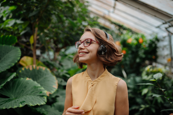 Beautiful woman standing in botanical garden, in the middle of lush green foliage with headphones on head, listening and enjoying music.