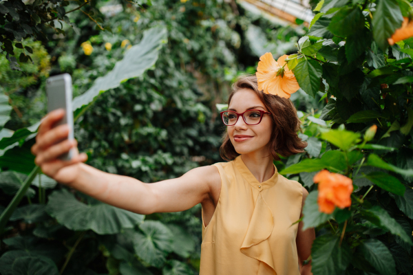 Beautiful woman taking selfie in botanical garden, holding smartphone, Woman standing in the middle of lush green foliage