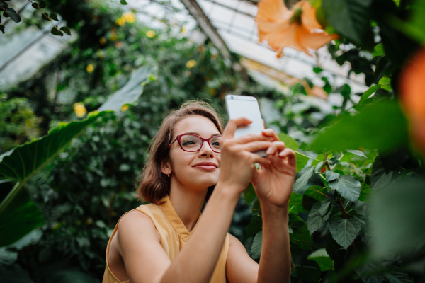 Woman in botanical garden, in the middle of lush green foliage, holding smartphone, taking picture of flower.